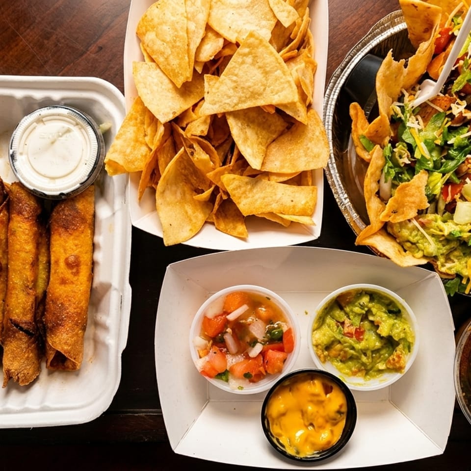 Assortment of Mexican takeout food including taquitos, chips, taco salad, guacamole, salsa, and queso.